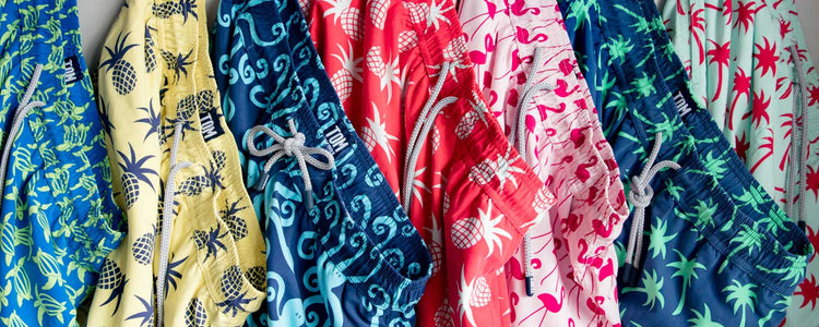 matching swimming trunks and clothing from Tom and Teddy showing a full range of swim shorts hangin up in different bright colours and designs from turtles, pineapples, octopus, flamingo, palm trees and more, perfect for father and son at the beach.